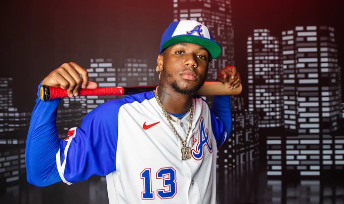 Ronald Acuña Jr. poses in the Braves' City Connect Uniforms with a bat behind his shoulders. The uniforms consist of a blue hat with a white panel, a white jersey with blue sleeves with "The A" on the right chest, and the player's number on the lower left side of the jersey. The pants are white. 