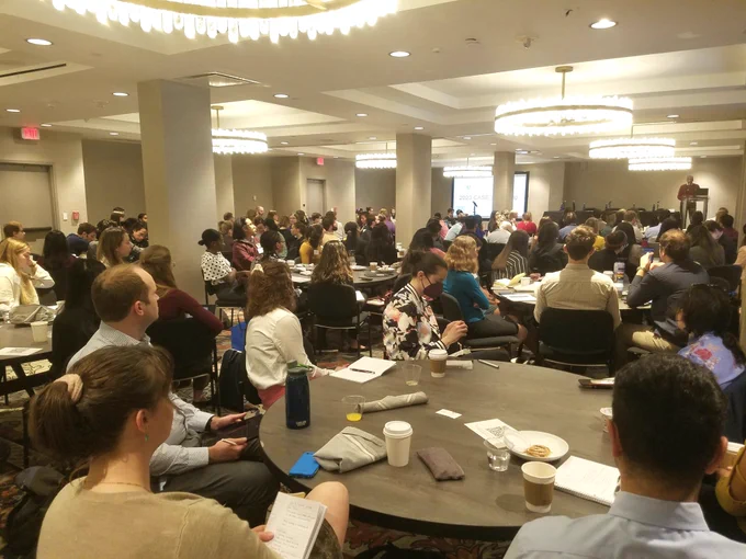A conference room full of people seated at round tables watch a speaker at the front of the room.