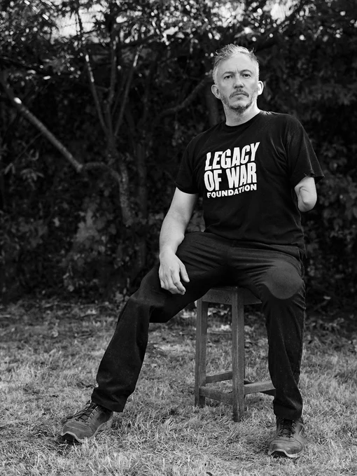 A black and white photograph of a man wearing a t-shirt that says “Legacy of War Foundation” sits on a stool outside. Image courtesy of Giles Duley.