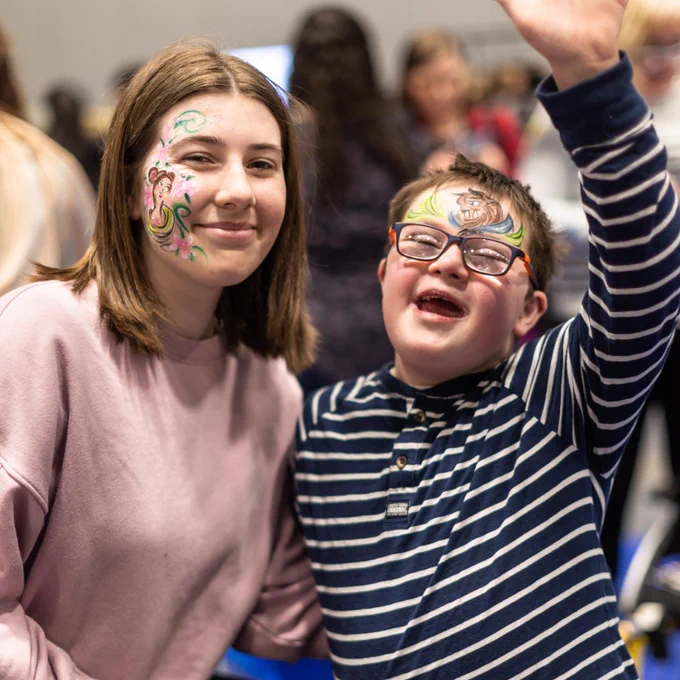 A young boy and girl are looking directly into the camera smiling. The boy is holding his hand up in the air and has a face painting of the beast from Beauty and the Beast on his forehead. The girl has Belle on the side of her face. 