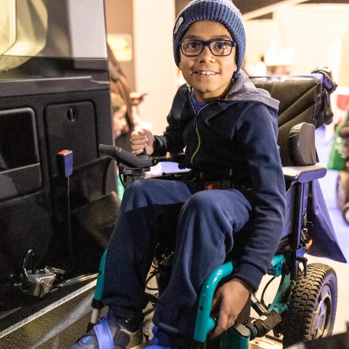 A young boy is seen in a wheelchair trialling one of the adaptable cars. He is wearing blue and is in a blue wheelchair. He is looking at the camera and smiling. 
