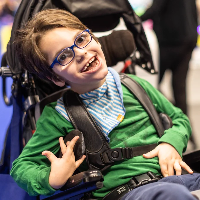 A young boy is sat in his wheelchair wearing a green top and blue and white bib. He is grinning with his mouth open and looking into the camera. 