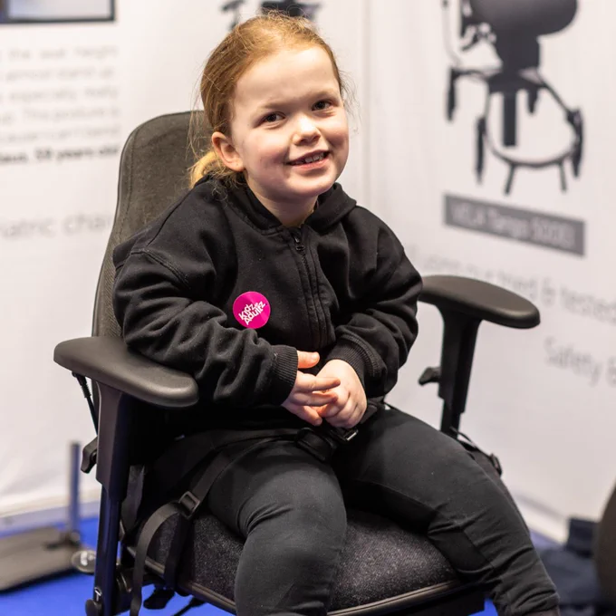 A young girl is sat in an adaptable chair. She is wearing all black and has a pink Kidz sticker on her hoodie. She is looking directly at the camera and smiling. 