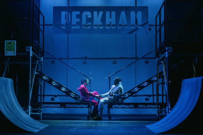 Two performers sit holding hands on stairs at the centre of a blue lit stage with Peckham in a cut out sign hanging above them and bike ramps either side of the stage 