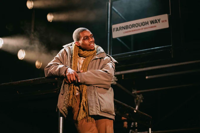 A young performer on stage leaning against a platform smiling in front of a road sign reading farnborough way and some stage lighting and haze  