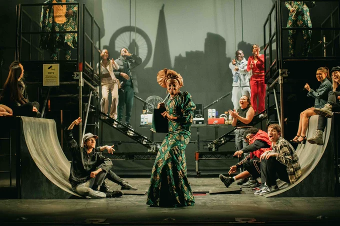 A female performers in a colourul green patterned dress dances at the front of a stage with bike ramps either side and platforms at the back filled with young performers smiling pointing at her