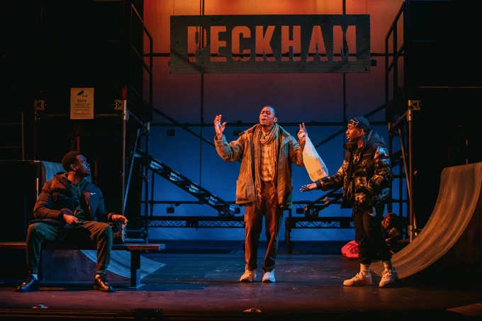 Three young performers in an animated conversation on a warmly lit stage, two wearing sportswear, the other with his arms raised carrying a plastic bag 