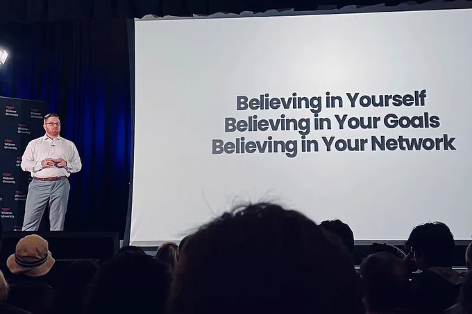 Ryan Raiker stands on stage next to a screen that says "Believing in yourself. Believing in your goals. Believing in your network."
