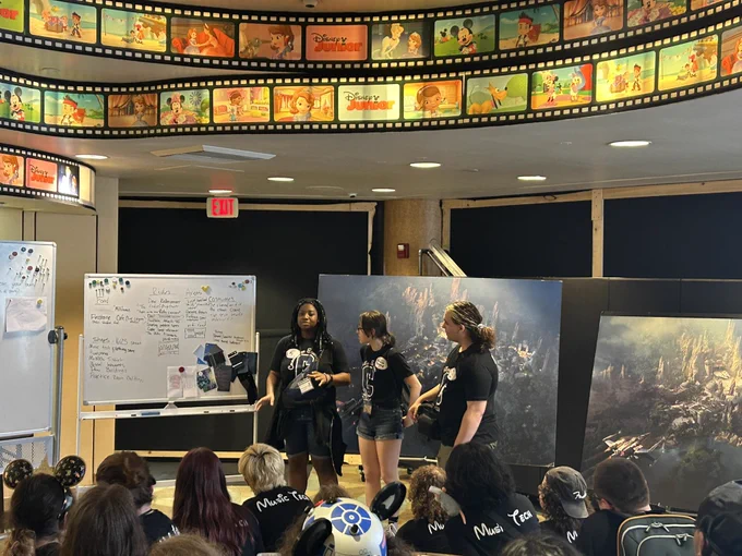 Three students presenting in front of a white board and large paintings underneath of a film strip boarder ceiling.