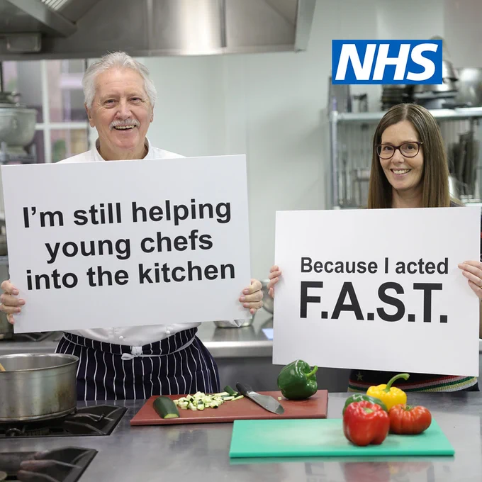 A man (Brian) and woman (Louise) stand inside a kitchen. Brian holds a sign that reads "I'm still helping young chefs into the kitchen". Louise holds a sign that reads "Because I acted F.A.S.T."