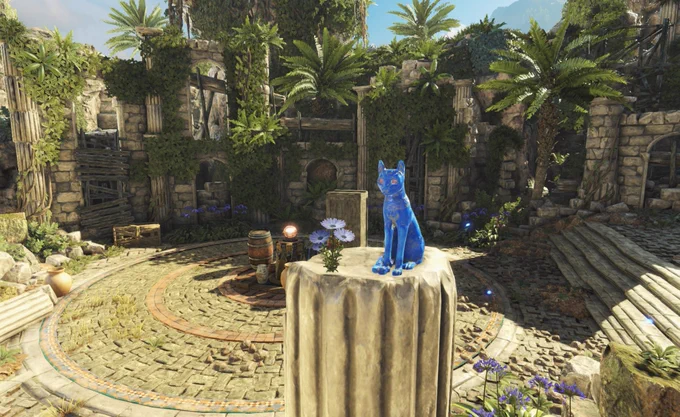 A view of a temple ruin overgrown with greenery and littered with a few barrels and boxes. A pillar stands slightly right aligned in the foreground with 3 purple flowers. To the right of these flowers sits one of Strange Brigade's famous Blue Cat idols. 