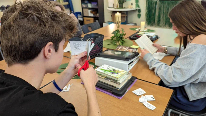 Student reading a book while another cuts out a small square of paper to place on a map