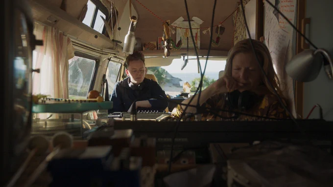 Image credit: Film still, Dream Agency, 2022

Alt text: A film still shows a man and a woman sat in a van, operating large audio decks, with a beach visible through the front window.
