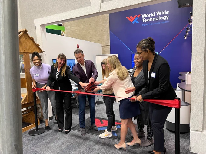 6 WWT employees stand in a line in front of a large red ribbon. In the center of the line, WWT Co-Founder and CEO, Jim Kavanuagh (left) and WWT EVP of Marketing, Sara Goellner help a young girl with large scissors to cut the ribbon. In the background is a large banner with a blue background and at the top, the WWT Logo sits between a blue and red gradient line. 