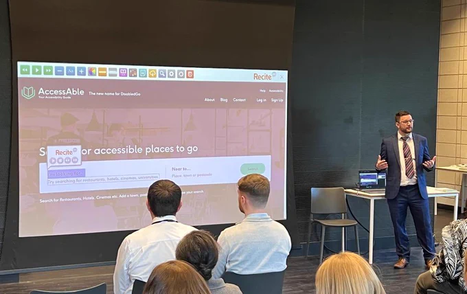 Dean Eales in front of an audience conducting a presentation on AccessAble. On display in the background is AccessAble’s homepage www.AccessAble.co.uk