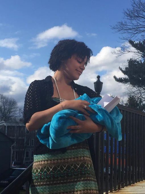 Moiya holding her printed out thesis wrapped in a blue blanket, smiling down at it lovingly. She is leaning against a black railing and there’s a big blue sky with some white clouds in the background