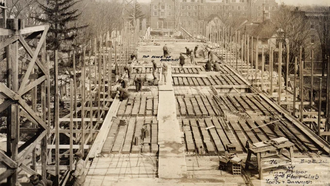 Workers build the structure of the Lawyer's Club dormitory in 1924. A scaffolding structure of wood supports the building. Around them, you can see the University of Michigan campus. "Top Floor of Dormitory" is written in the center.
