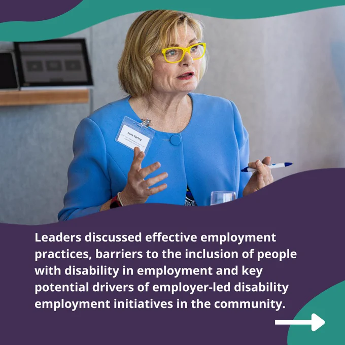 A woman is sitting in a room and talking to a group of people. She has mid length blond hair and is wearing yellow framed glasses and a blue jacket. Text: Leaders discussed effective employment practices, barriers to the inclusion of people with disability in employment and key potential drivers of employer-led disability employment initiatives in the community. 