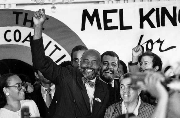 Mel King, wearing a suit, stands with his hand held high in victory. Behind him supporters cheer and there is a sign that says Mel King For Mayor