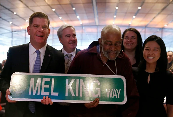 Mayor Marty Walsh and Mel King hold a large street sign that says "Mel King Way." On the left, Mayor Michelle Wu smiles at the camera. More smiling people are behind them. 