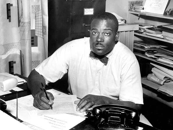 Mel King sits at a desk. He's obviously very busy as there is a lot of paper on his desk and he is hard at work. He's wearing a short sleeved white shirt and a bow tie. 