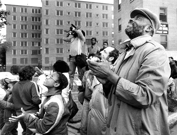 Mel King is in the foreground stands looking up, clapping. He is bearded, wearing a trench coat and a dapper hat. Behind him is a crowd, news photographers, and in the distance, apartment buildings. 