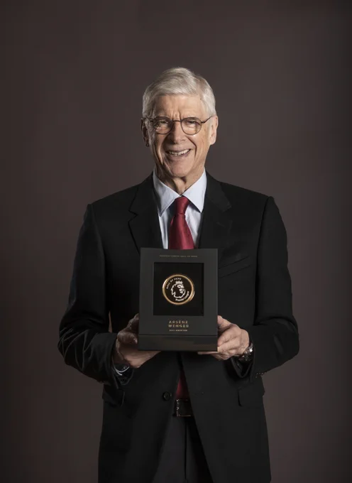 Arsène Wenger, dressed in a suit, smiles into the camera while holding up his Premier League Hall of Fame award