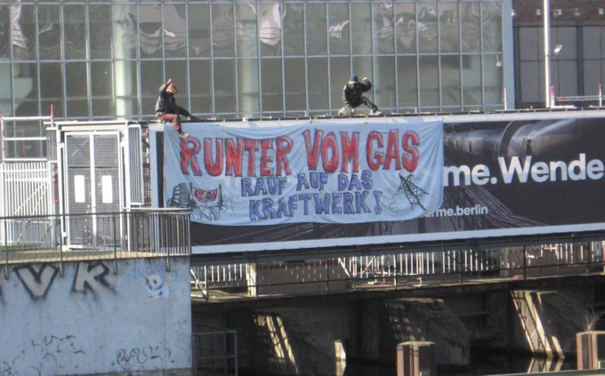 2 Menschen sitzen auf dem Gelender einer Brücke vom Vattenfall-Werk in Berlin über ein ausgerolltes Banner. 
Da steht:
Runter vom Gas rauf auf das Kraftwerk.