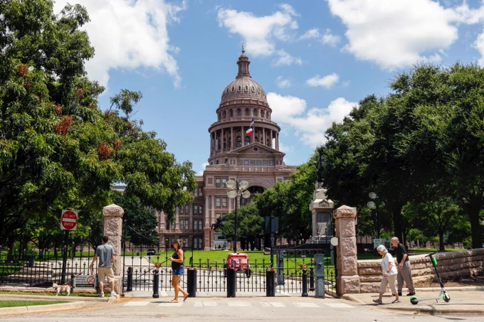 The photo shows the Texas Capitol during the daytime as depicted by the bright blue sky and the sunlight hitting the sidewalk next to the building. Pedestrians are casually walking past the building. Photo by Jamie Hwang.