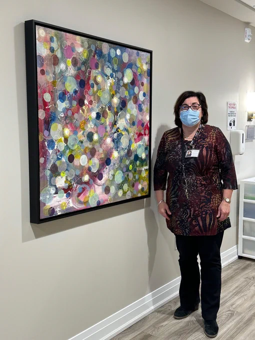 Hospice Vaughan Executive Director Stephanie Karapita, masked, poses with an Artworks for Cancer artwork installed in the north wing corridor of Hospice Vaughan: Denise Morrison, Forever Blowing Bubbles, acrylic on canvas