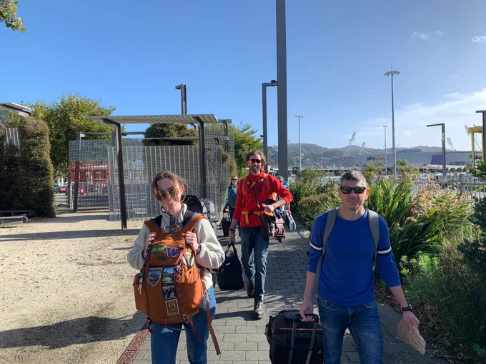 Members of the research cruise smiling and walking towards the camera on a sunny day outdoors. They all have their shades on and are carrying their packed luggage and bags.