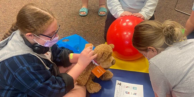 A young child sits on the floor with a teddy bear talking to Child Life specialists. 