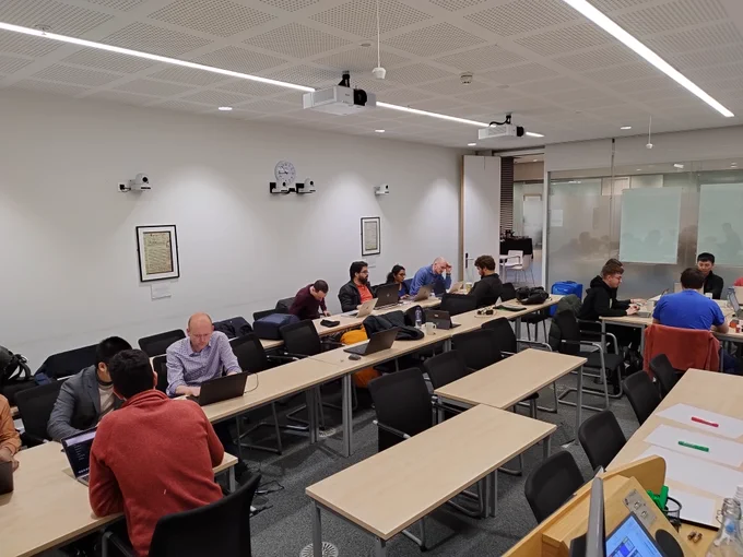 Three of the groups working in a lecture room. Each group is sitting around a table and writing code together.