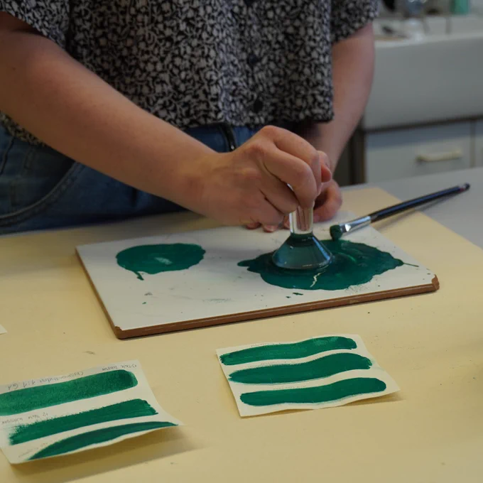Detail of a person preparing paint by grinding pigment and binding media. The main focus is a hand using a glass muller to grind the green pigment on a white plate. In front of the plate there are several pieces of paper on which the green paint has been applied. 