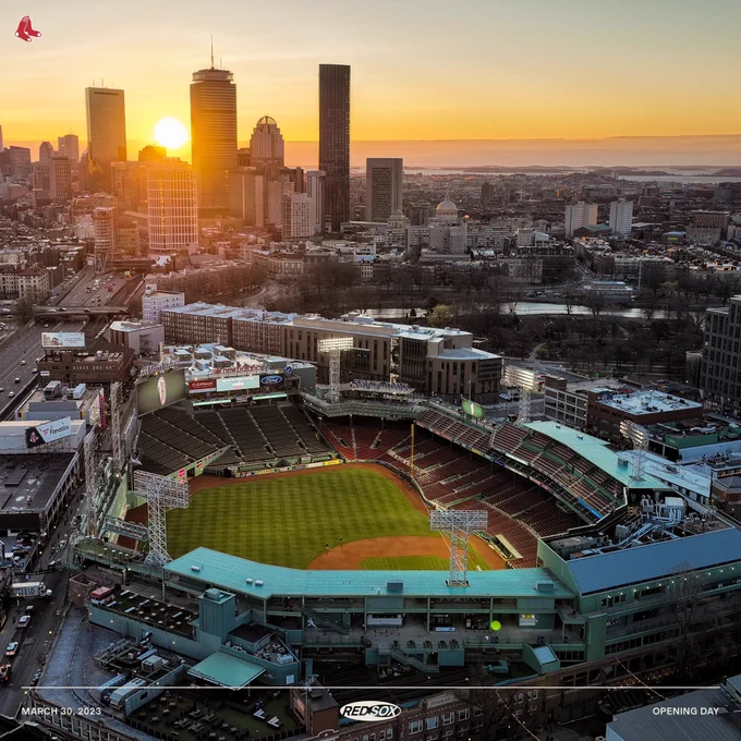 A wide and high shot of an empty Fenway with green grass and lights on as the sun rises along the Boston skyline on Opening Day.