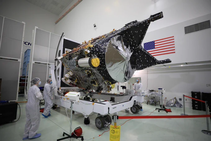 A team of engineers in full coveralls, hairnets, and masks look at the Psyche satellite in a clean room. The satellite is about the size of a golf cart and is mounted on a turnable structure.
