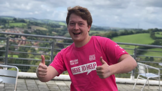 A student wearing a bright pink top that says "HERE TO HELP" written on the front. The student is smiling with their mouth open, and has both thumbs up.