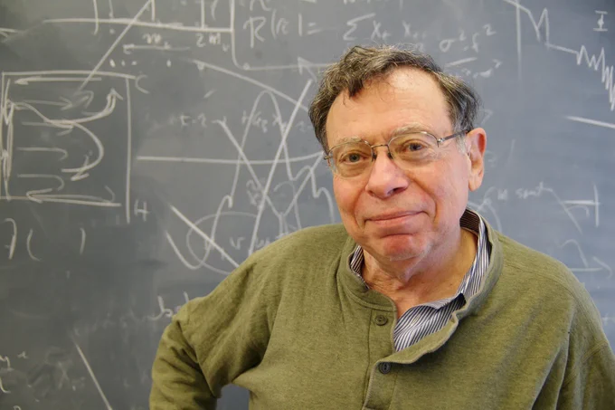 A portrait of Carl Wunsch smiling at the camera in front of a blackboard