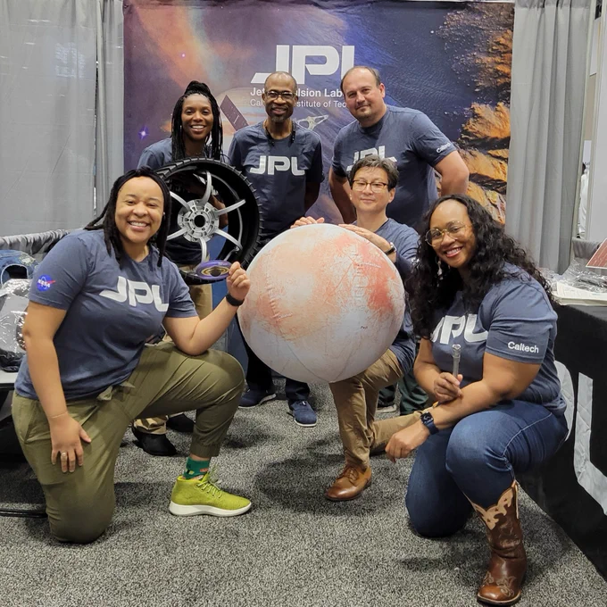 The is a group photo of JPL staff at the booth at the NSBE conference. All six are wearing JPL shirts and smiling at the camera. 