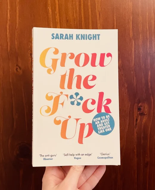 I'm holding up a copy of Grow the F*ck Up by Sarah Knight in my left hand in front of a timber background.