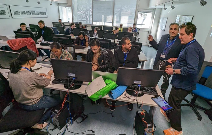Classroom setting, with large windows in the background. Several people work on computers. They are the participants at the Joint ICTP-IAEA Advanced School/Workshop on Machine Learning in Citizen Science, held in Trieste from 27 February to 3 March 2023. 
App Inventor education specialist Angie Zhou (bottom left, sitting) and Robert Parks (far right, standing) instruct the 19 students from 16 countries attending the workshop.
(Photo: App Inventor Foundation)