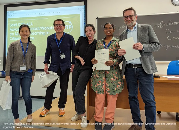 3 women and 2 men, in a lecture room, standing and smiling to the camera. Some hold certificates. They are among the participants, lecturers, organizers of the Joint ICTP-IAEA Advanced School/Workshop on Machine Learning in Citizen Science, held in Trieste from 27 February to 3 March 2023. The name of the workshop is also reflected on the screen behind them.
(Photo: App Inventor Foundation)