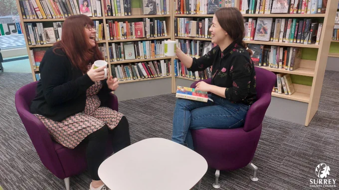 Two women sit in their library, holding Reading Friends mugs. They are chatting and laughing over a copy of Lessons in Chemistry by Bonnie Garmus. In the bottom right corner, the white Surrey County Council logo. 