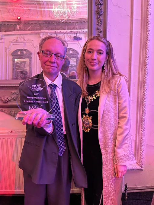 Roy is wearing a grey suit with a blue patterned tie, and holding a glass award with Wolfgang's name on it. Emma, the mayor, is stood to his right, wearing a long cream patterned coat with her mayoral civic regalia around her neck.