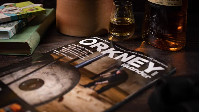 Image of the Orkney Islander magazine on a table, with Orkney guide books and a bottle and glass of Highland Park whisky.