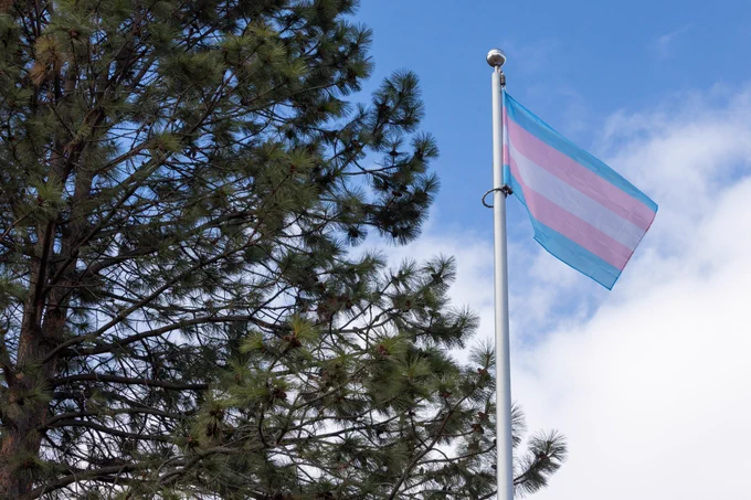 A photo of a transgender flag flying at TRU. There is some blue sky with white fluffy clouds and a tree in the background.