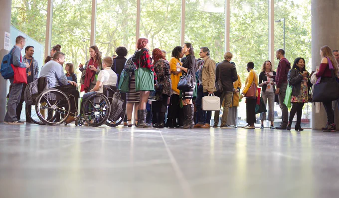 Group of diverse people mingling: 2 people in wheelchairs, various ethnicities, various genders, some casually dressed, some in a suit.