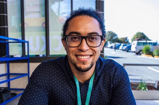 Adam Fryer is a young Black man with long hair pulled back in a ponytail. He is outdoors, with a building an parking lot behind him. He wears glasses, has a goatee, and has on a navy shirt with small white polka dots.