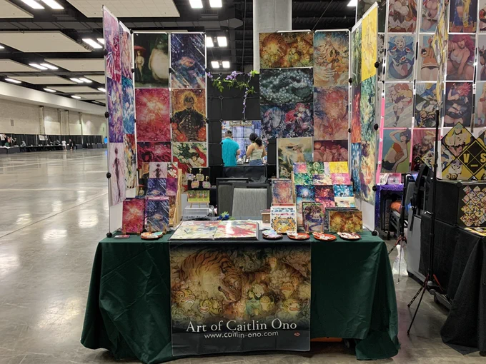 Artist alley table at Kawaii Kon for Art of Caitlin Ono. A table filled with art, covered with a green tablecloth and a banner with the Paper Tigers art piece labeled “Art of Caitlin Ono” www.caitlin-ono.com.