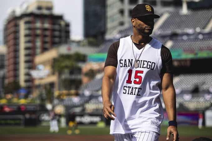 Photo of Xander Bogaerts on the field during BP. He is wearing a brown Padres hat and a brown t-shirt with a white San Diego State basketball jersey with the number 15 on it over top of the t-shirt.